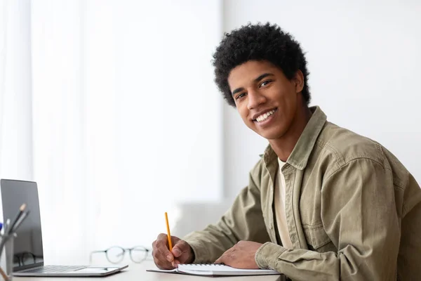 Portrait of positive black teenage student with laptop and notebook looking at camera and smiling at home, copy space