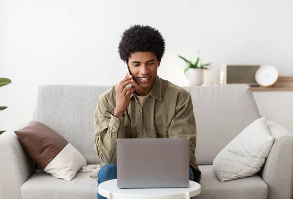 Conceito de e-learning. Funky estudante negro do sexo masculino se comunicando no celular na frente do computador portátil em casa — Fotografia de Stock
