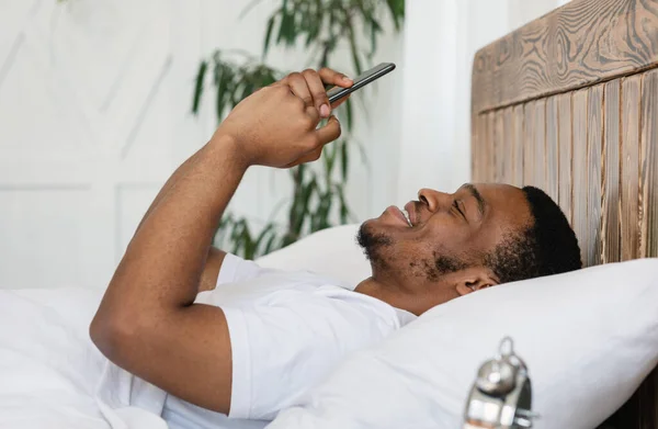 Alegre afro-americano cara usando smartphone deitado na cama dentro de casa — Fotografia de Stock