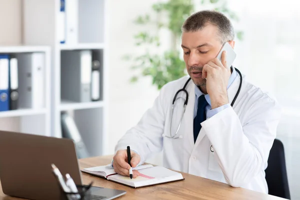 Lekarz rozmawia przez telefon z pacjentem i pisze notatki. — Zdjęcie stockowe