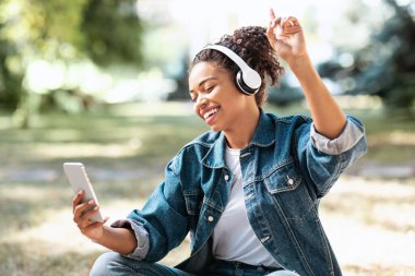 Neşeli siyah kadın kulaklıklı, müzik dinleyen açık havada oturan.