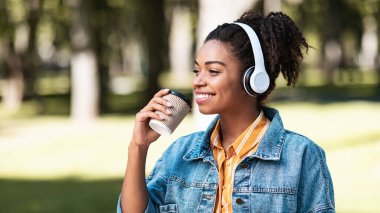 Siyah kadın kahve içiyor, parkta müzik dinliyor.