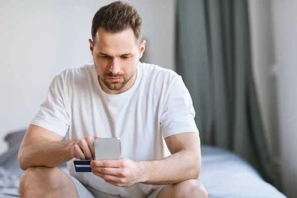 Hombre sosteniendo Smartphone y tarjeta de crédito sentado en el dormitorio — Foto de Stock