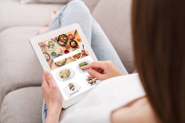 Evde yemek sipariş etmek için modern uygulama. Kanepede rahat bir şekilde oturan ve tabletle yemek seçen bir kadın.