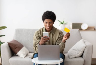 Bilgisayarlı havalı Afro-Amerikalı genç cep telefonu ve kredi kartını evde online alışveriş için kullanıyor.