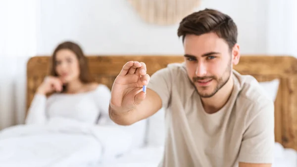Si concentri su pillola azzurra in mani di giovane uomo felice, e la moglie sorridente si siede in letto in interno moderno di camera da letto — Foto Stock