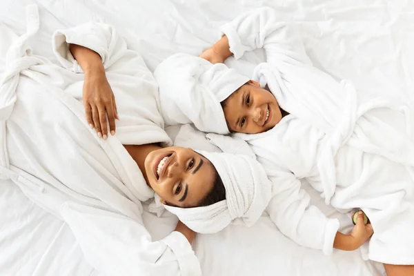 Χαρούμενη Αφρικανή μητέρα και κόρη με μπουρνούζια και πετσέτες ξαπλωμένες στο κρεβάτι — Φωτογραφία Αρχείου