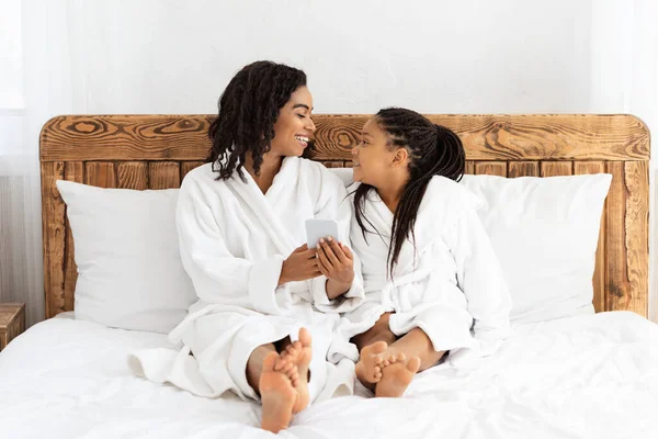 Mamá africana y su hija usando Smartphone mientras se relaja en la cama en batas de baño — Foto de Stock