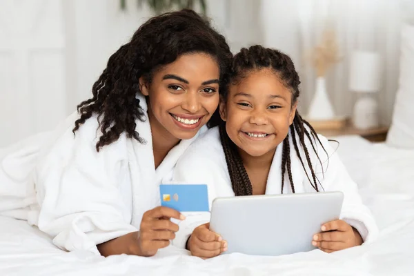 Black Mother And Daughter In Bathrobes Posing With Digital Tablet And Credit Card