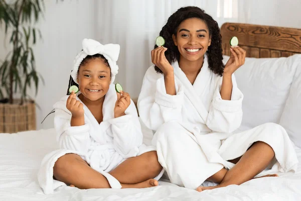Smiling African Mom And Daughter Sitting In Bathrobes Holding Cucumber Slices
