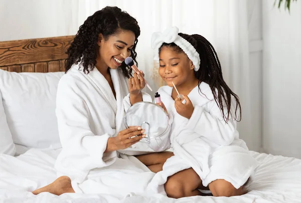 Bela mãe negra feliz e filha pequena em roupões de banho aplicando maquiagem juntos — Fotografia de Stock