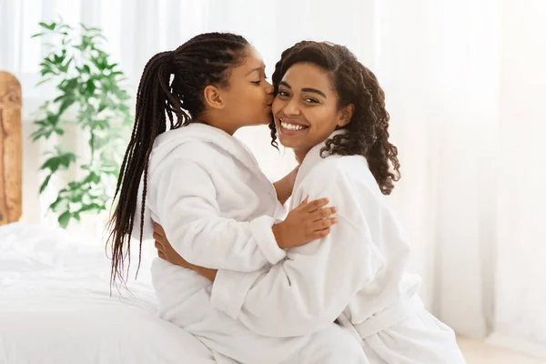 Día de las Madres. Adorable niña negra besando a mamá en casa, usando albornoces — Foto de Stock