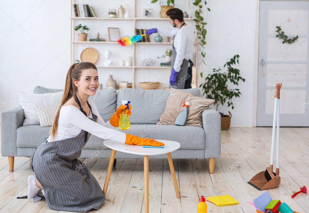 Smiling woman in apron and rubber gloves with spray wipes dust from table, husband in shelves