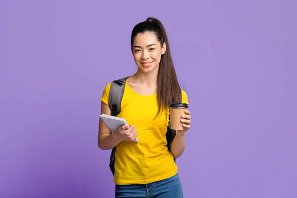 Jovem estudante asiática feliz com mochila e café sobre fundo roxo — Fotografia de Stock