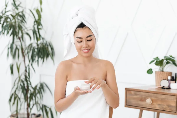 Sorrindo asiático jovem senhora envolto em toalha usando creme — Fotografia de Stock