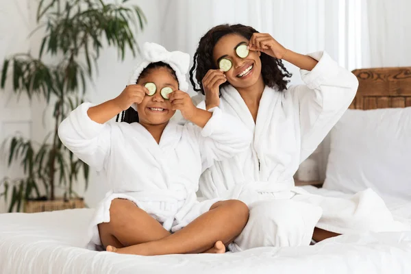 Giocoso nero madre e figlia in accappatoi applicando pezzi di cetriolo agli occhi — Foto Stock