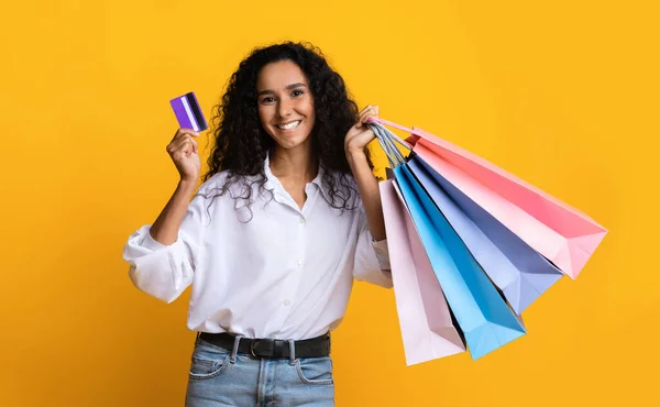 Felice donna bruna con borse della spesa e carta di credito su sfondo giallo — Foto Stock