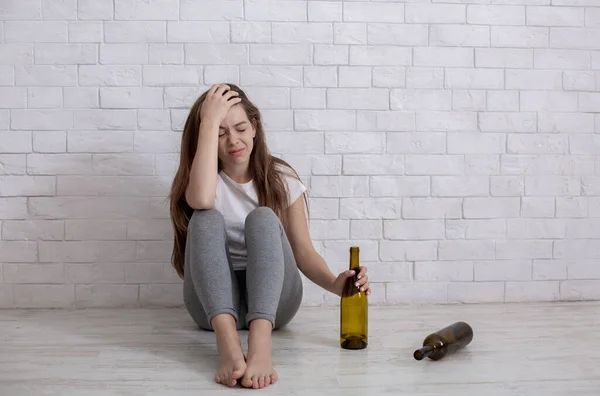 Концепція зловживання алкоголем. Молода пригнічена жінка з пляшкою бузку сидить на підлозі вдома, порожній простір — стокове фото