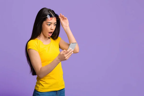 Geschokt aziatische vrouw op zoek naar smartphone scherm met open mond, paarse achtergrond — Stockfoto