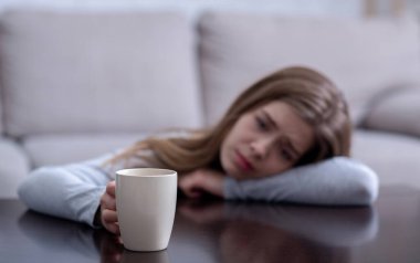 Üzgün genç kadın masaya yaslanıyor ve elinde sıcak içecekle depresyondan muzdarip bir fincan tutuyor. Seçici odaklanma. Boşluğu kopyala