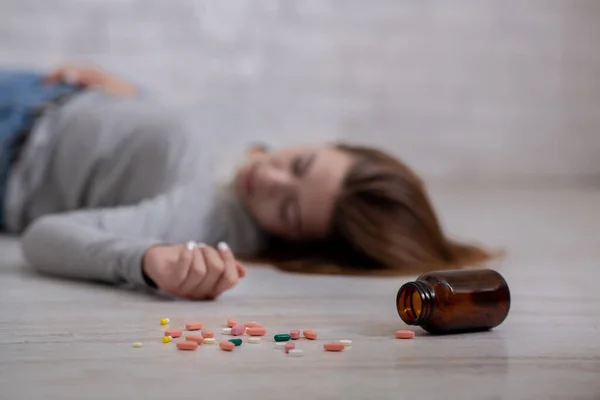 Mujer joven acostada en el suelo con una botella de tabletas esparcidas por el suelo, suicidándose, enfoque selectivo — Foto de Stock