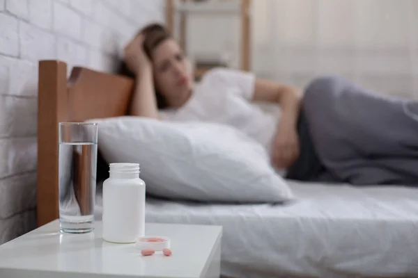 Frasco de comprimidos e vidro de água na mesa de cabeceira e jovem deprimida deitada na cama no fundo, espaço de cópia — Fotografia de Stock