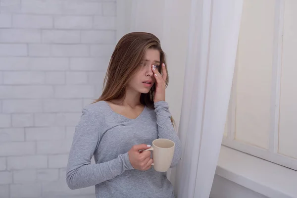 不快乐的年轻女士站在窗边，边喝咖啡边哭，在屋里 — 图库照片
