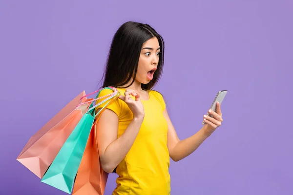 Onlineförsäljning. Chockad asiatisk kvinna tittar på smartphone och hålla Shopping väskor — Stockfoto