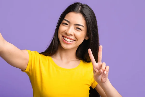 Zorgeloze jonge aziatische vrouw nemen selfie en tonen vrede gebaar — Stockfoto