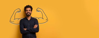 Hintli adam kollarını açık sarı bir arkaplan önünde kavuşturarak güvenle ayakta duruyor. Kol kasları onun gücünü, gücünü ve dayanıklılığını canlı bir ortamda gösteriyor..