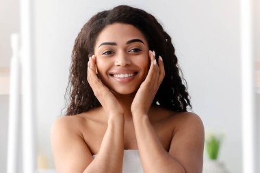 Mükemmel sonuç, rutin prosedürler, cilt nemlendirici, güzellik bakımı, covid-19. Mutlu bin yıllık Afro-Amerikan kadın beyaz havluyla yüzüne dokunuyor, yatak odasının içindeki büyük aynaya bakıyor.