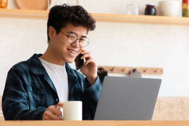 Evde kendini izole etme, kovid 19 karantina, uzaktan çalışma, müşteri araması. Sosyal mesafeler için modern aletler. Gözlüklü gülümseyen Asyalı çocuk dizüstü bilgisayara bakıyor ve mutfakta telefonla konuşuyor.