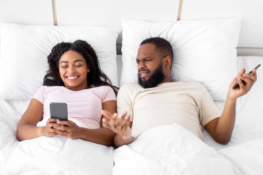 Displeased young african american guy gesturing, spreads hands, look at woman smartphone on bed in bedroom interior. App and online shopping, addiction to technology, online games and social networks