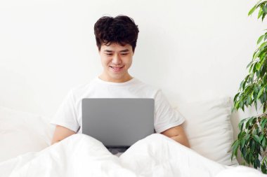 Yatak odasında iş, sosyal ağlar, sörf, internette gezinme, modem cihazı. Gülümseyen Asyalı adam uyandı, beyaz yatağa oturdu, iç dizüstü bilgisayarda çalıştı, arka planda ışık duvarı, serbest alan