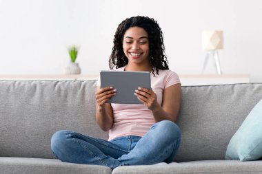 Modern teknoloji, yeni normal, covid-19 salgını sırasında sosyal mesafeler. Gülümseyen bin yıllık sevimli Afro-Amerikan kadın tablete bakıyor ve az gri oturma odasının içinde, boş bir alanda komik videolar izliyor.