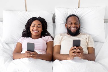 Smiling young african american husband and wife typing on phones, playing online games on bed in bedroom interior, top view. Sleeplessness, addiction to gadgets and social networks, new app and ad