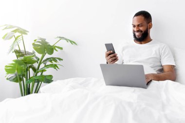Beyaz tişörtlü mutlu siyah sakallı adam yatağında dizüstü bilgisayarda çalışıyor, yatak odasının içinde telefonla mesajlar okuyor. Covid-19 salgını sırasında evde uzak iş ve serbest çalışma için modern cihazlar
