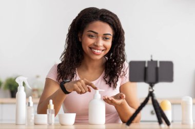 Modern güzellik blogcusu sosyal ağlar için video dersi veriyor. Neşeli Afro-Amerikan bayan tripodda akıllı telefondan webcam 'e bak, nemlendirici kullan ve iç mekândaki kozmetik ürünlerini incele.