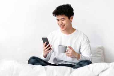 Covid-19 salgını, sabah haberleri, kahve ve hafta sonları boş vakitlerde evde kalın. Milenyum Asyalı pijamalı bir adamın elinde akıllı telefon ve yatak odasının içinde bir fincan sıcak içecek tutmasına sevindim.