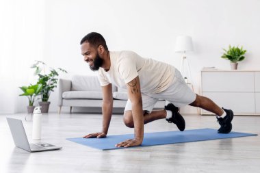 Beyaz spor giysili, sakallı, mutlu siyah erkek oturma odasında dizüstü bilgisayarda bacak egzersizi yapıyor ve video seyrediyor. Çevrimiçi sınıf spor, ders ve eğitim covid-19 karantinası sırasında
