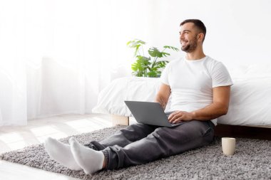 Gülümseyen milenyum kaslı Avrupalı erkek yatak odasının iç kısımlarında fikir üretiyor ve laptop üzerinde çalışıyor, boşluğa bakın. Ev işleri, modern teknoloji ve sosyal mesafeler, sabahları alet edevat ve sohbet.
