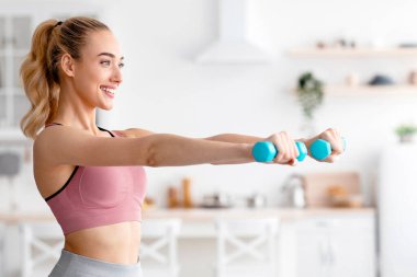 Aktif spor, spor salonunda beden eğitimi, kovid 19 karantinası. Spor giyimli genç ve çekici sarışın kadın halterleri kaldırıyor, minimalist mutfak iç kısımlarında kol egzersizleri yapıyor.