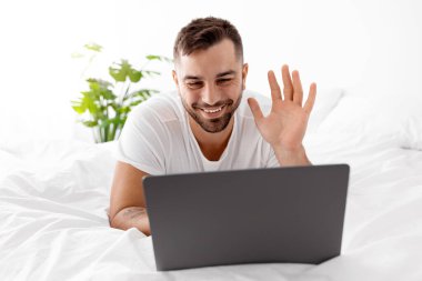 Gülümseyen yakışıklı milenyum kaslı beyaz adam bilgisayarda yazı yazıyor, dizüstü bilgisayarında el sallıyor yatak odasındaki yatak odasında web kamerasıyla yatıyor. Görüşme kumandası, video çağrısı, sosyal uzaklık ve sohbet için aygıt
