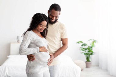 Gülümseyen genç Afro-Amerikan kocası hamile karısına sarılıyor, minimalist yatak odasında karnına dokunuyor. Bebek beklentisi, ilişki ve aşk, ortaklık, mutlu aile ve gelecekteki ebeveynlik