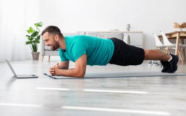 Sakallı, genç ve yakışıklı bir sporcu oturma odasının içinde düzgün bir ders veriyor. Spor kumandası, vücut bakımı, covid-19 salgını sırasında koç ve çevrimiçi sınıfla egzersizler, profil