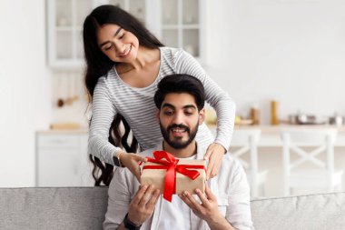 Evde tatil, yıldönümü, doğum günü ve covid-19 salgını sırasında sevgililer günü. Gülümseyen genç, güzel bir Arap kadın kocasına hediye verir. Oturma odasında kutlama yapar.