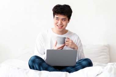 İş sırasında evde kahve molası, sıcak içecek ve aletlerle dinlenme. Beyaz tişörtlü gülümseyen Asyalı genç adam yatak odasında iç tarafta dizüstü bilgisayarla oturuyor, bardağı tutuyor ve boş alana bakıyor.