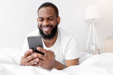 Mutlu yıllar Afro-Amerikan sakallı adam yatak odasının içinde beyaz yatakta uzanıyor ve akıllı telefonuyla daktilo yazıyor. Covid-19 tecrit, sosyal uzaklık, yeni uygulamayla sohbet, reklam, teklif ve indirim