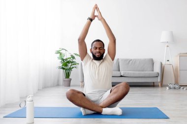 Sakallı, yerde nilüfer pozisyonunda oturan ve oturma odasında yoga yapan sakallı, Afro-Amerikan genç adam. Spor, sağlıklı yaşam tarzı, karantina sırasında evde çalışma.