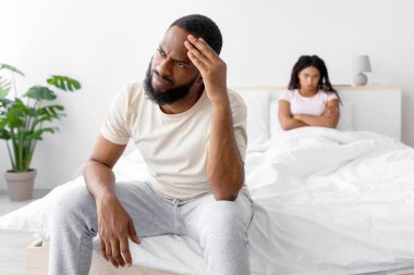 Unhappy sad young african american male ignores female in comfortable bed in bedroom interior, empty space. Emotions after quarrels and problems at home, facial expressions during covid-19 pandemic